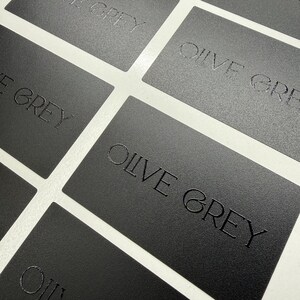 May include: A collection of rectangular matte black business cards arranged in a grid pattern. Each card features the text OLIVE GREY embossed in a clean, sans-serif font. The surface of the cards has a fine, textured finish throughout.