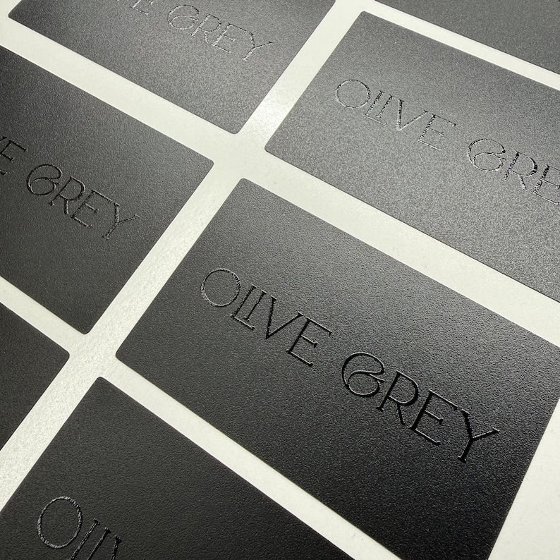 May include: A collection of rectangular matte black business cards arranged in a grid pattern. Each card features the text OLIVE GREY embossed in a clean, sans-serif font. The surface of the cards has a fine, textured finish throughout.