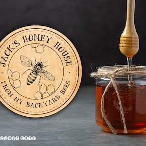 Peut inclure: Une &eacute;tiquette ronde brune avec le texte "Jack's Honey House From My Backyard Bees" en lettres noires. Une illustration en noir et blanc d'une abeille avec du miel est au centre de l'&eacute;tiquette. Un pot de miel avec un n&oelig;ud en ficelle est en arri&egrave;re-plan.