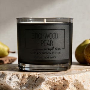 May include: A clear glass candle jar with a matte black label featuring the text BIRCHWOOD + PEAR, coconut wax, HAND POURED IN THE US, and NET WT 8OZ (237G). The candle has a white wax fill and a single centered wick, presented in a cylindrical vessel.