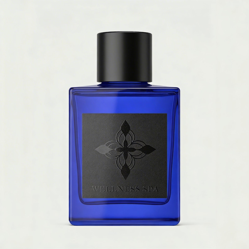 May include: A rectangular cobalt blue glass bottle with a matte black cylindrical cap. A square black label on the front features a dark embossed floral emblem above the text WELLNESS SPA in a simple serif font, centered on the glass surface.