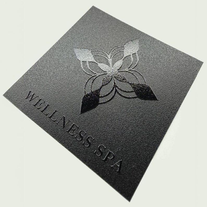 May include: A square black card with a textured surface featuring the text WELLNESS SPA in embossed capital letters. Above the text is a symmetrical, four-pointed geometric emblem with a gradient finish that transitions from light to dark.