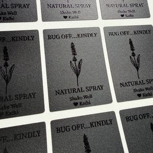 May include: A grid of rectangular black labels featuring a line drawing of a lavender sprig. The text reads BUG OFF...KINDLY, NATURAL SPRAY, Shake Well, and a heart symbol followed by the name Kathi, all printed in a dark, textured finish.