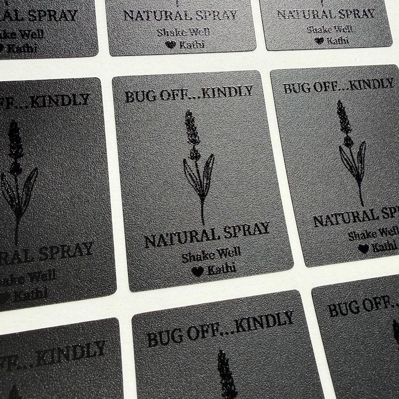 May include: A grid of rectangular black labels featuring a line drawing of a lavender sprig. The text reads BUG OFF...KINDLY, NATURAL SPRAY, Shake Well, and a heart symbol followed by the name Kathi, all printed in a dark, textured finish.