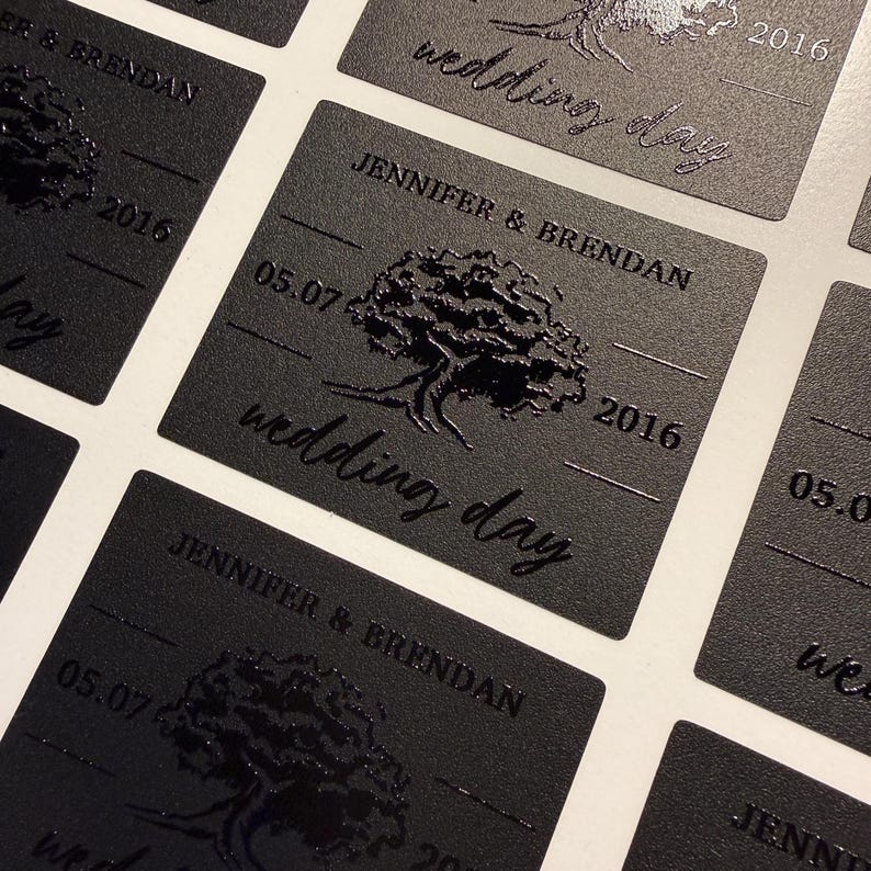 May include: A series of rectangular black labels featuring a glossy embossed tree graphic in the center. The text reads JENNIFER & BRENDAN, 05.07, 2016, and wedding day. The labels have a matte finish with raised, shiny black lettering and imagery.