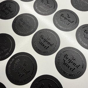 May include: A collection of circular black stickers featuring the embossed text So Beyond Sweet and the website URL WWW.SOBEYONDSWEET.COM arranged in a repeating pattern. Each sticker has a matte finish with the lettering slightly raised.