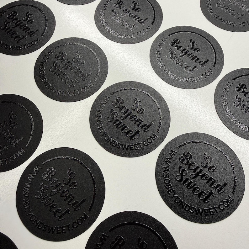 May include: A collection of circular black stickers featuring the embossed text So Beyond Sweet and the website URL WWW.SOBEYONDSWEET.COM arranged in a repeating pattern. Each sticker has a matte finish with the lettering slightly raised.