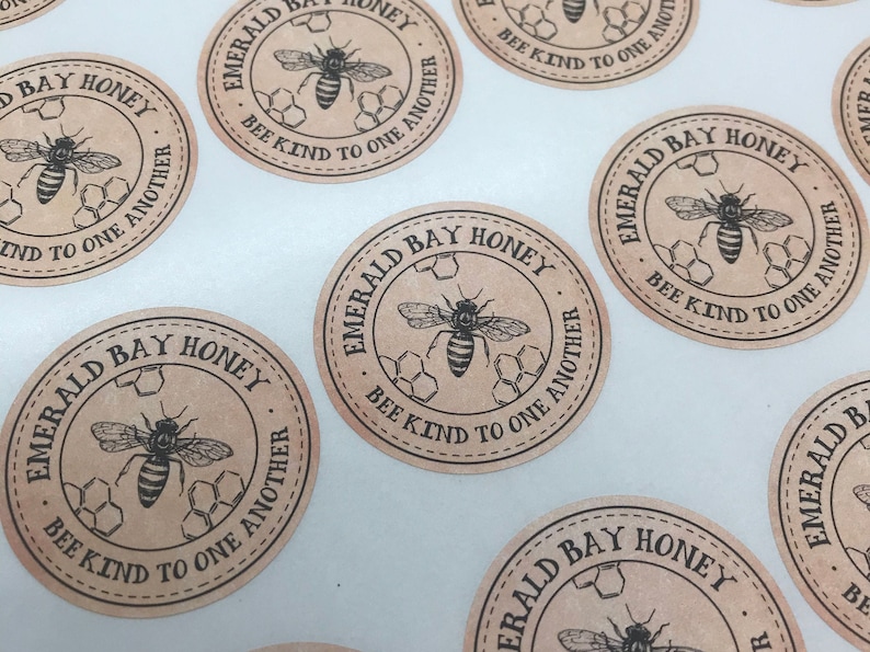 Peut inclure: Une collection d'autocollants ronds en papier brun avec une illustration en noir et blanc d'une abeille et d'un nid d'abeilles. Le texte sur les autocollants est "Emerald Bay Honey" et "Bee Kind to One Another".