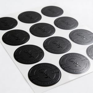 May include: A sheet of circular black stickers featuring an embossed design of a central flower. Each sticker includes the text NATURAL BEAUTY, ESTD, 2026, and YOUR SLOGAN HERE arranged within a circular border surrounding the floral illustration.