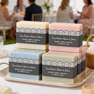 May include: A collection of rectangular soap bars in cream, pink, blue, and green, each wrapped in a dark gray paper band featuring a white lace pattern and the printed text: From Denise's Shower to Yours, DENISE'S BRIDAL SHOWER, OCTOBER 18, 2025.