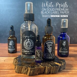 May include: Five glass bottles with black labels and white text. The labels feature illustrations of flowers, a woman's face, and a leaf. The bottles contain various liquids, including a clear spray bottle, a brown dropper bottle, a blue dropper bottle, and two smaller dropper bottles. The bottles are arranged on a wooden surface. The text on the labels reads "Company Name", "Eden Spa", "Custom Text", "4 oz | 118 ml", "MOOD DROPS", "GLOW DROPS", "Burdock Root", "1 fl. oz (30 ml)", "Soul Say", and "One Trace Charity Mist Set with Love".