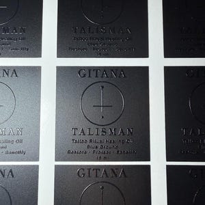 May include: A grid of square black labels with embossed text and graphics. Each label features the word GITANA at the top, a circular symbol with a vertical line, and the text TALISMAN Tattoo Ritual Healing Oil Blue Ground Restore Protect Sanctify 15 ml.