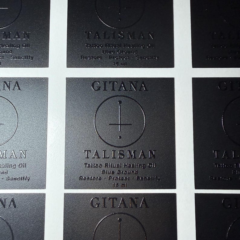 May include: A grid of square black labels with embossed text and graphics. Each label features the word GITANA at the top, a circular symbol with a vertical line, and the text TALISMAN Tattoo Ritual Healing Oil Blue Ground Restore Protect Sanctify 15 ml.