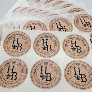 Peut inclure: Gros plan sur dix &eacute;tiquettes rondes en papier brun avec le texte "Herrington Brothers Honey" et un graphique d'abeille noire. Les &eacute;tiquettes sont imprim&eacute;es avec le texte "Virginia, Georgia - Local Raw Honey".
