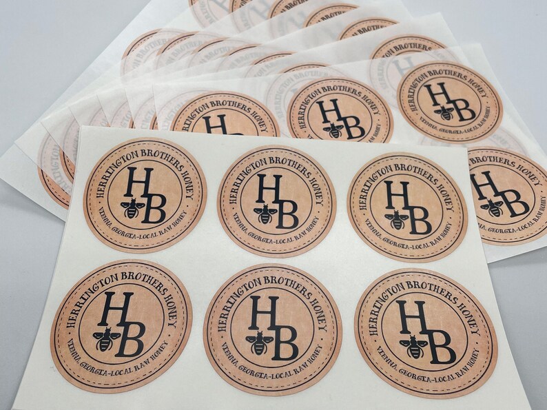 Peut inclure: Gros plan sur dix &eacute;tiquettes rondes en papier brun avec le texte "Herrington Brothers Honey" et un graphique d'abeille noire. Les &eacute;tiquettes sont imprim&eacute;es avec le texte "Virginia, Georgia - Local Raw Honey".