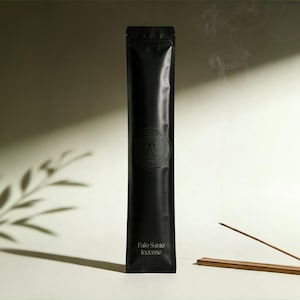 May include: A tall, rectangular black pouch containing Palo Santo incense. The packaging features a circular logo with the letters WMA and the text Palo Santo Incense printed in white at the bottom. A single incense stick rests on a wooden holder.