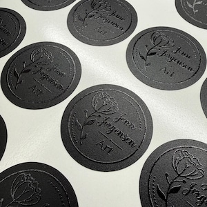 May include: A collection of circular black stickers featuring a debossed floral design and the text Jenn Jorgensen Art. Each sticker has a dotted border surrounding a single flower illustration and the brand name printed in a clear, legible font.