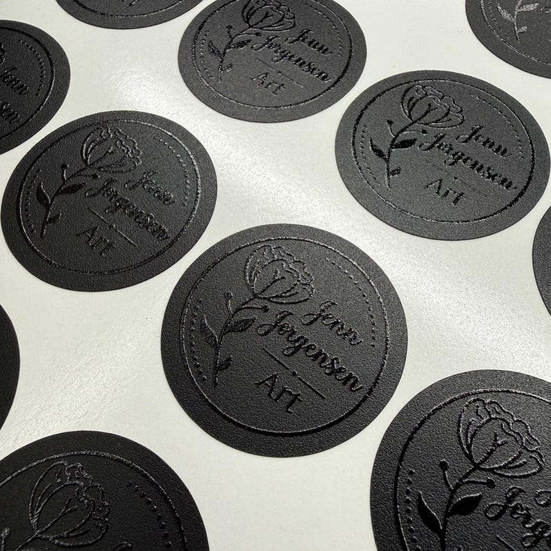 May include: A collection of circular black stickers featuring a debossed floral design and the text Jenn Jorgensen Art. Each sticker has a dotted border surrounding a single flower illustration and the brand name printed in a clear, legible font.