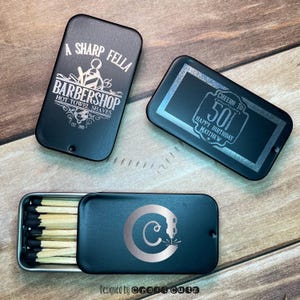 May include: Three black metal matchboxes with engraved designs. The first box has the text "A Sharp Fella Barbershop Hot Towel Shaves Est. 2019". The second box has the text "Cheers to 50 Happy Birthday Matthew". The third box has the letter "C" with a stylized design.