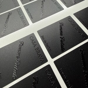 May include: A collection of rectangular black labels with a textured finish. Each label features the embossed text "CASA BRAVA" in a sans-serif font, "Femme Fatale" in a cursive script, and "Eau de Parfum" in a smaller, clean typeface.