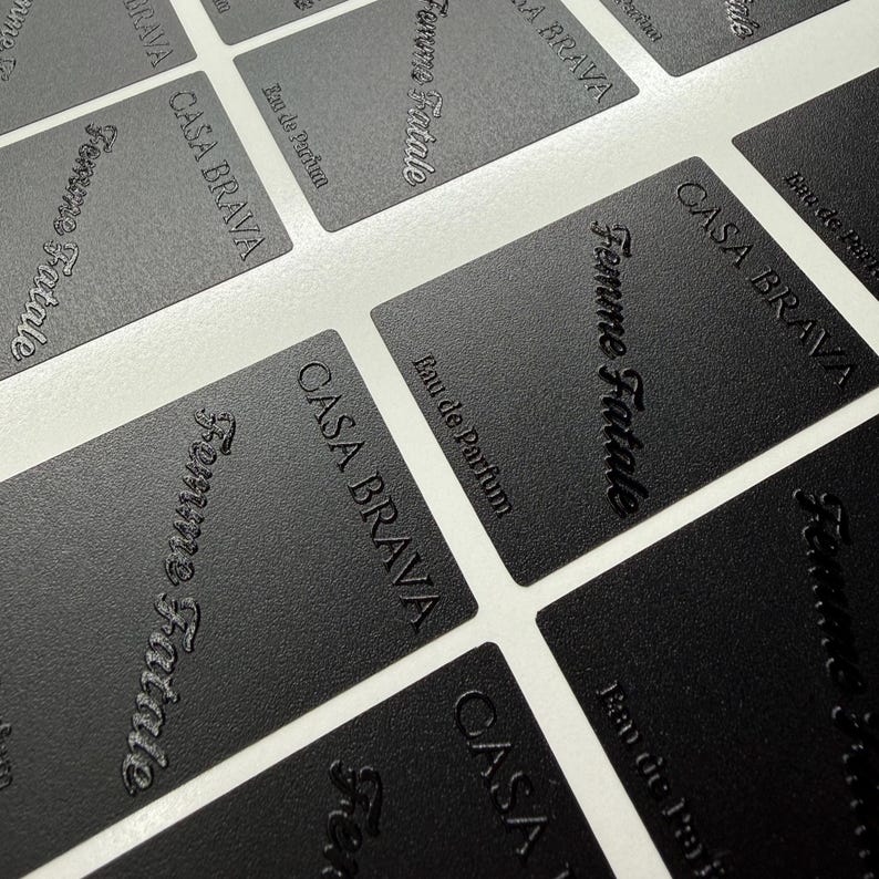 May include: A collection of rectangular black labels with a textured finish. Each label features the embossed text "CASA BRAVA" in a sans-serif font, "Femme Fatale" in a cursive script, and "Eau de Parfum" in a smaller, clean typeface.