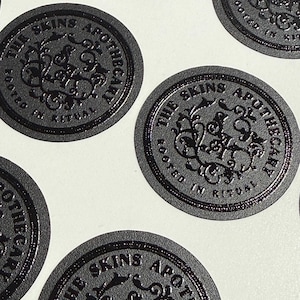 May include: A collection of circular black stickers featuring a raised, embossed design. Each sticker displays the text THE SKINS APOTHECARY and ROOTED IN RITUAL surrounding a central ornate, swirling monogram logo with intricate botanical-style details.