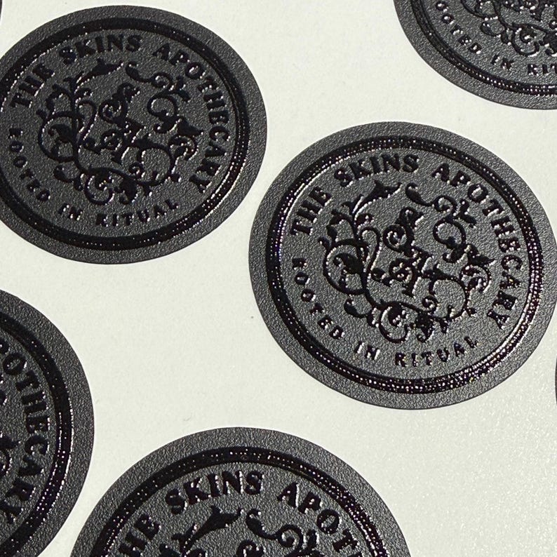 May include: A collection of circular black stickers featuring a raised, embossed design. Each sticker displays the text THE SKINS APOTHECARY and ROOTED IN RITUAL surrounding a central ornate, swirling monogram logo with intricate botanical-style details.
