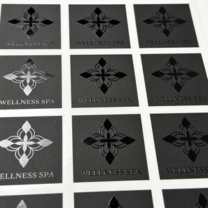 May include: A sheet of square stickers featuring a black background with a central symmetrical floral emblem and the text WELLNESS SPA printed below. Some stickers display the design in a matte black finish, while others feature a reflective silver foil.