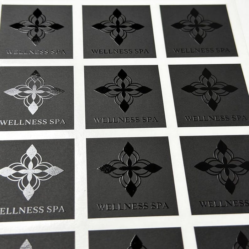 May include: A sheet of square stickers featuring a black background with a central symmetrical floral emblem and the text WELLNESS SPA printed below. Some stickers display the design in a matte black finish, while others feature a reflective silver foil.