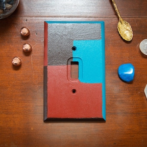 Puede incluir: Una cubierta de interruptor de luz rectangular con bloques de color rojo, gris y turquesa. La cubierta tiene una abertura para el interruptor y dos orificios para tornillos. La imagen también incluye piedras decorativas, una cuchara dorada y objetos de color cobre.
