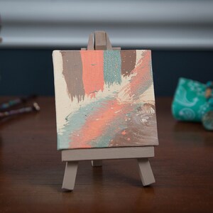 Puede incluir: Una pequeña pintura abstracta sobre lienzo con tonos marrones, rosados y azules. La pintura está en un pequeño caballete.