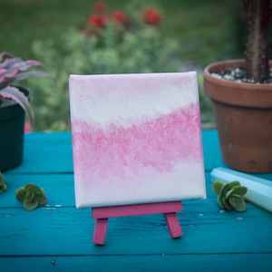 Puede incluir: Una pequeña pintura de acuarela rosa y blanca sobre lienzo, expuesta en un caballete rosa. La pintura está sobre una mesa de madera azul con suculentas verdes.