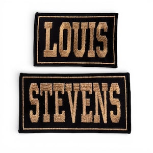 May include: Two rectangular black patches with gold embroidered text. The top patch reads "LOUIS", and the bottom patch reads "STEVENS". The patches have a gold border and are likely for clothing or accessories.