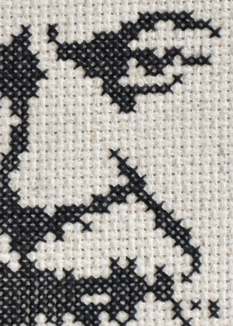 May include: Black and white cross-stitch embroidery on a beige fabric background. The design features a stylised floral pattern.