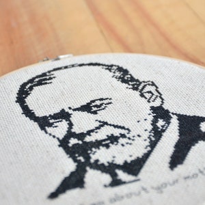 May include: Black and white cross-stitch embroidery of a man's face with a beard. The embroidery is in a round wooden hoop and the text "Tell me about your mother" is stitched below the face.