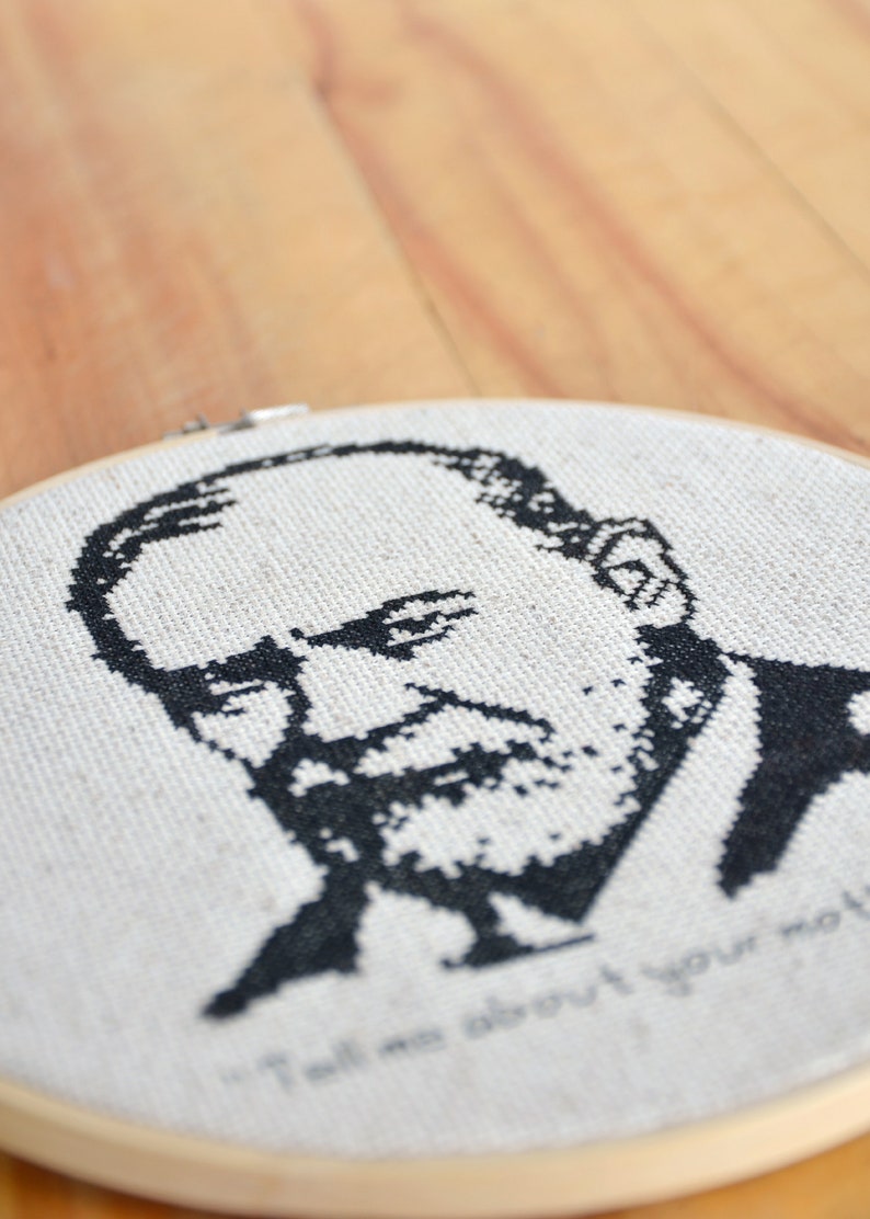 May include: Black and white cross-stitch embroidery of a man's face with a beard. The embroidery is in a round wooden hoop and the text "Tell me about your mother" is stitched below the face.