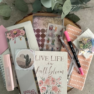 May include: A collection of stationery supplies including a pink highlighter, a black pen, a pink and white polka dot zippered pouch, a notebook with the text "Live Life in Full Bloom" and a notebook with the text "Spread Kindness".