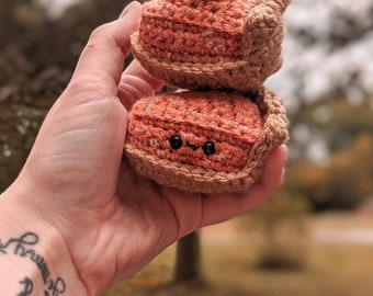 May include: Two crocheted pumpkin pie plush toys stacked on top of each other. The top pie has a white crocheted dollop of whipped cream on top. The bottom pie has a cute face with black eyes.