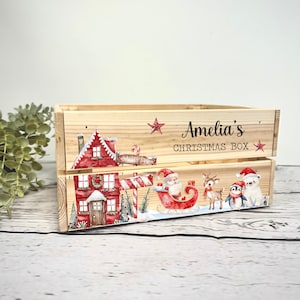 May include: A rectangular wooden crate featuring a watercolor illustration of a red house, Santa in a sleigh, a reindeer, a penguin, and a polar bear. The text "Amelia's CHRISTMAS BOX" is printed in black on the upper right side of the crate.
