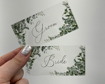 May include: Two clear acrylic signs with a green eucalyptus leaf design. The top sign says "Groom" and the bottom sign says "Bride".