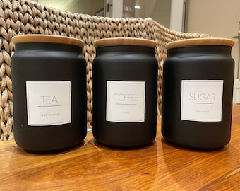 May include: Three black canisters with bamboo lids and white labels. The labels read "TEA english breakfast", "COFFEE instant", and "SUGAR granulated".