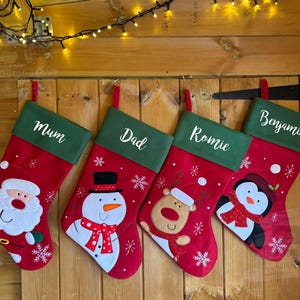 May include: Four red Christmas stockings with green felt cuffs and personalised names: Mum, Dad, Romie, and Benjamin. Each stocking features a different embroidered character: Santa Claus, a snowman, a reindeer, and a penguin.