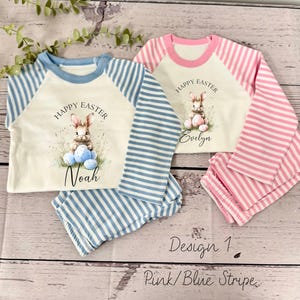 May include: Two pairs of pajamas for children, one in pink and white stripes and one in blue and white stripes. Both pajamas feature a white shirt with a bunny holding Easter eggs and the text "Happy Easter". The pink pajamas have the name "Evelyn" on them and the blue pajamas have the name "Noah" on them. The text "Design 1 Pink/Blue Stripe" is written below the pajamas.