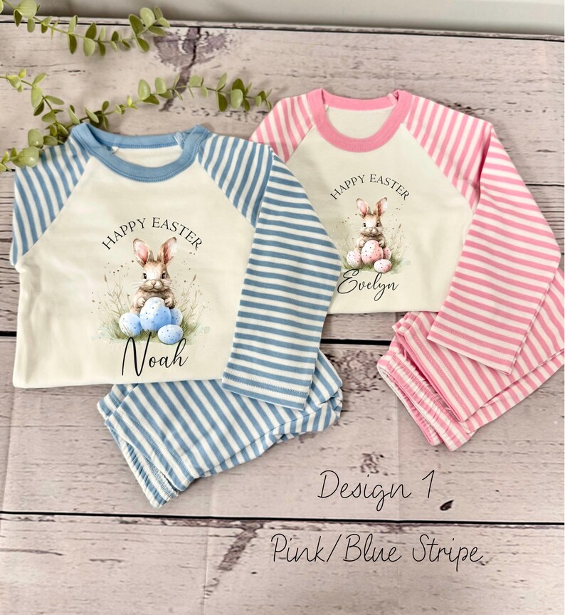 May include: Two pairs of pajamas for children, one in pink and white stripes and one in blue and white stripes. Both pajamas feature a white shirt with a bunny holding Easter eggs and the text "Happy Easter". The pink pajamas have the name "Evelyn" on them and the blue pajamas have the name "Noah" on them. The text "Design 1 Pink/Blue Stripe" is written below the pajamas.