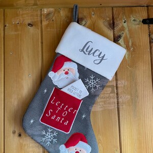 May include: Grey knitted Christmas stocking with a white cuff personalised with the name "Lucy". The stocking features a red and white Santa Claus face with a white beard and a red hat. The stocking also has a red pocket with the words "Letter to Santa" and a white note that says "Pocket to hold letters".