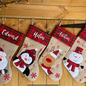 May include: Four Christmas stockings with names: Edward, Helen, Arthur, and Nora. Each stocking features a red tartan cuff and a festive character: Santa, penguin, reindeer, and snowman. They hang on a wooden door.
