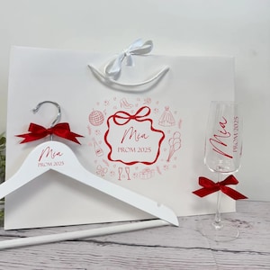 May include: A white gift bag, hanger, and champagne flute set for prom. The bag and flute are decorated with the name "Mia" and "Prom 2025" in red script. The hanger has a red bow. The bag has a red design with prom-related icons.