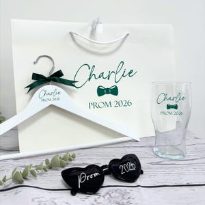 May include: A white wooden hanger with a green bow, a white gift bag, and a pint glass, all personalized with the name "Charlie" and "PROM 2026" in green. Black heart-shaped sunglasses with "Prom" and "2026" are also present.