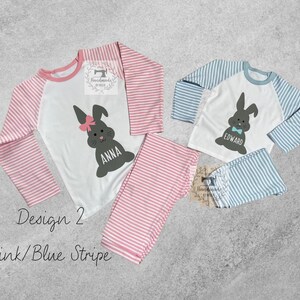 May include: Two children's pajamas with a pink and blue stripe design. The pajamas feature a white top with a bunny rabbit graphic and the names "Anna" and "Edward".