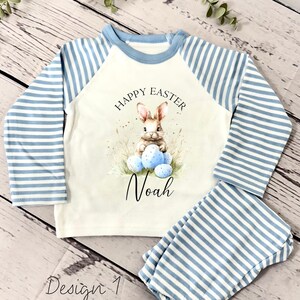 May include: A white long-sleeved shirt with blue and white striped sleeves. The shirt has a graphic of a bunny holding Easter eggs and the text "Happy Easter" and "Noah". The shirt is paired with blue and white striped pants.